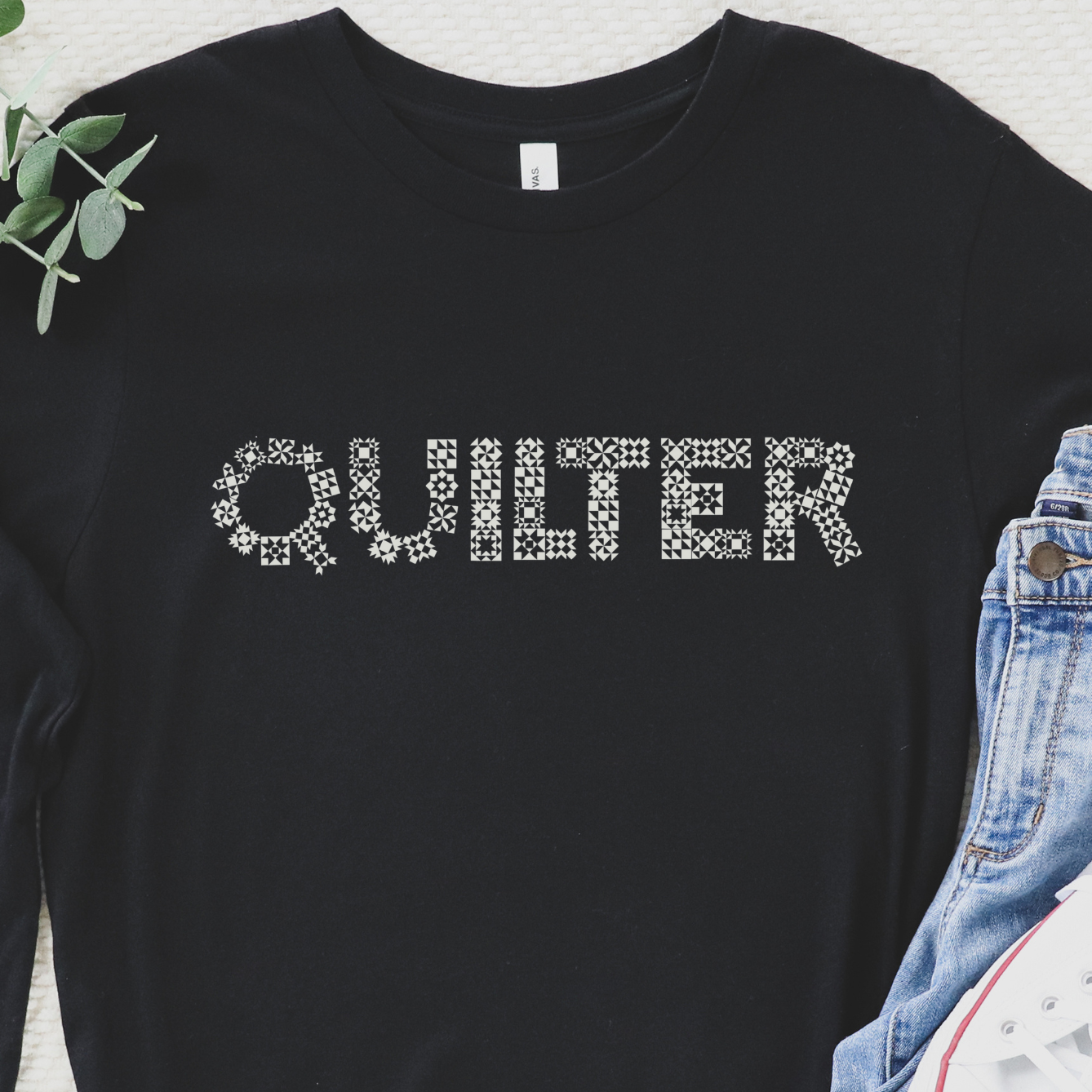 Black long-sleeve shirt with 'QUILTER' text, jeans, and white sneakers on a textured surface.